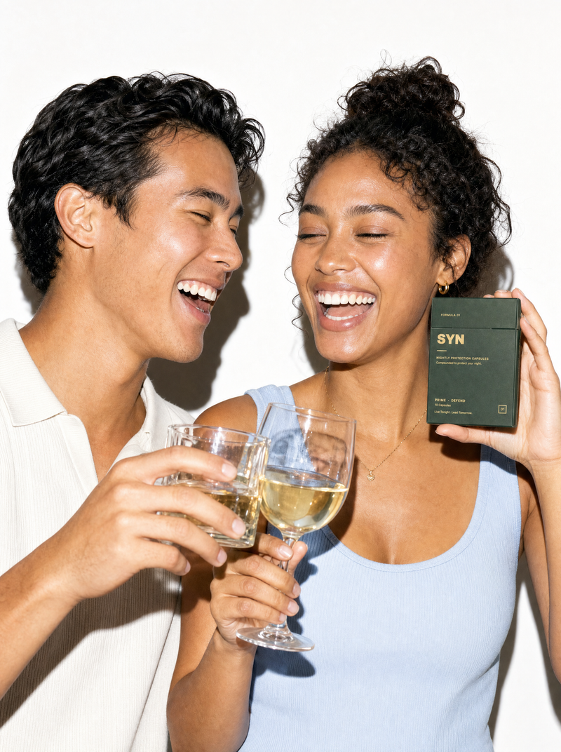 A couple laughing and toasting with white wine, one holding a box of SYN