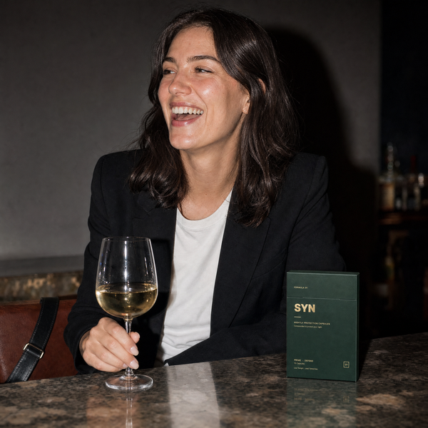 A woman laughing over white wine at a hotel bar, a box of SYN on the counter