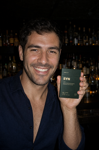 A man smiling at a cocktail bar, holding a box of SYN