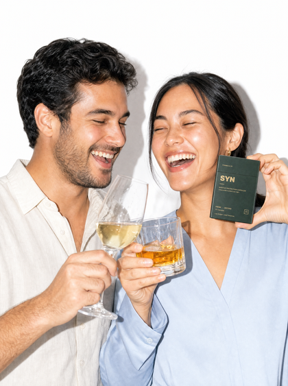A couple laughing and toasting with wine and whisky, holding a box of SYN